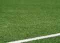 Campo de futebol de grande dimensão com grama sintética verde, marcação branca e alambrado ao fundo