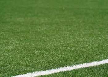 Campo de futebol de grande dimensão com grama sintética verde, marcação branca e alambrado ao fundo